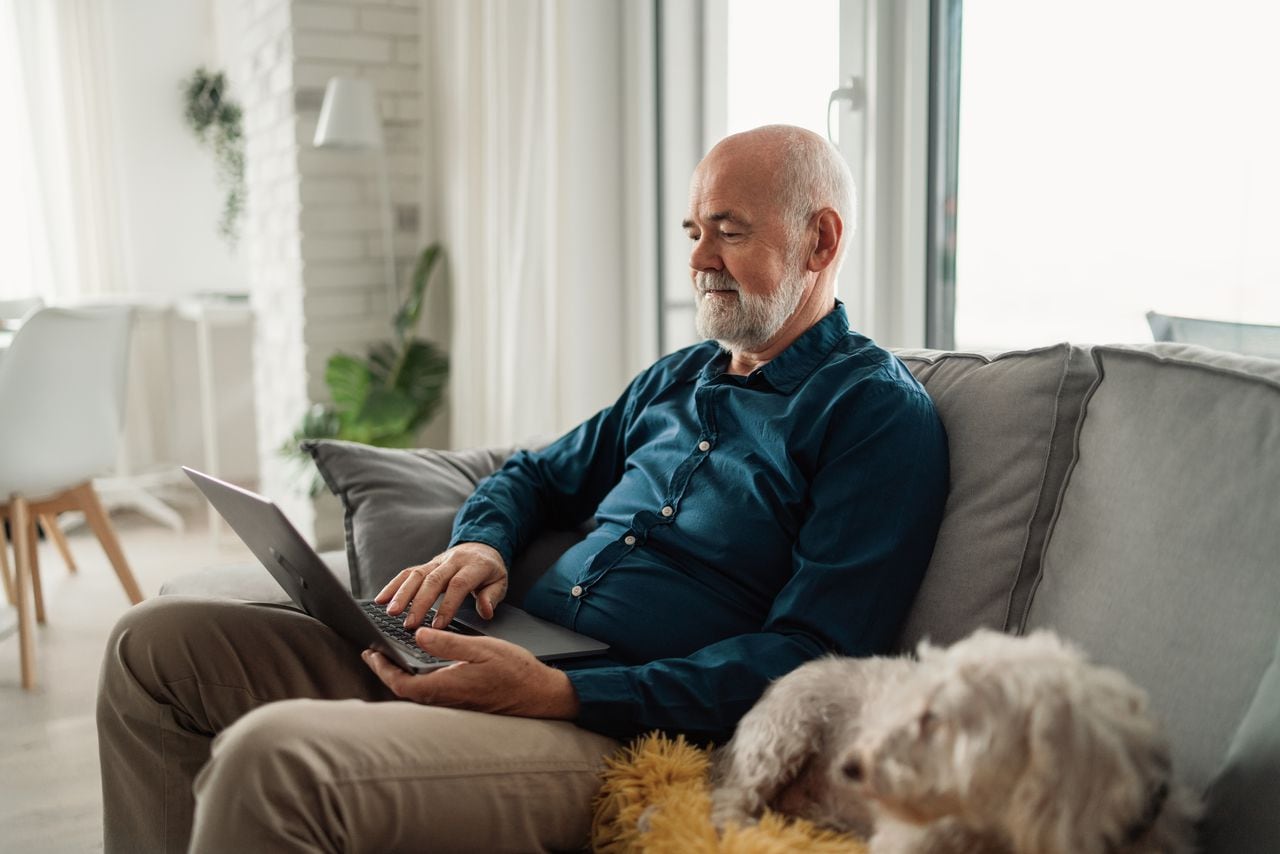 Senior man sitting on sofa with his dog and using laptop at home.